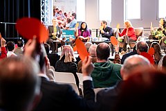 In einem Raum mit dunklen Seitenwänden und einer hellen Fensterfront hinter dem Podium sitzen verschiedene Menschen in Stuhlreihen und schauen auf die am Podium sitzenden Personen, die auf dem Foto im Hintergrund verschwommen sind. Die Menschen im Publikum halten runde grüne oder rote Zettel in die Höhe, um damit abzustimmen. 
