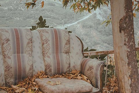 Ein vintage-Sofa in weiß, rosa und blau, steht in der Natur. rechts davon ein Baum. Die herbstlichen Blätter fallen auf die ERde und auf das Sofa. Im Hintergrund sind Berge zu sehen