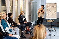 Personen sitzen gemeinsam, eine Frau steht vorne und erklärt etwas an einer Flip-Chart