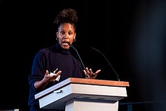 Auf einem Podium steht eine Schwarze Frau, hinter einem weißen Stehpult. Sie trägt einen schwarzen Rollkragenpullover. Hinter ihr steht ein weißer Aufsteller mit dem Logo des BBE (drei blaue Halbkreise mit der Abkürzung BBE in schwarz, darunter steht "Bundesnetzwerk Bürgerschaftliches Engagement") unter dem Logo steht entwickeln, vernetzen.