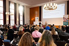 In einem Raum mit hohen Fenstern sitzen Personen in Stuhlreihen und schauen auf eine weiße Leinwand mit einer Power Point Präsentation.